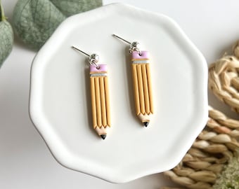 May include: A pair of yellow pencil earrings with pink eraser tops and silver earring hooks. The earrings are displayed on a white plate.