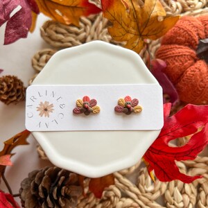May include: A pair of small, brown, orange, and red turkey-shaped earrings on a white card with the text "Carline & Co.". The earrings are displayed on a white saucer surrounded by fall-colored leaves.