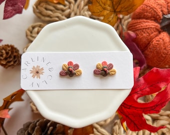 Puede incluir: Un par de pequeños pendientes en forma de pavo de color marrón, naranja y rojo sobre una tarjeta blanca con el texto "Carline & Co.". Los pendientes se muestran en un platillo blanco rodeado de hojas de otoño.