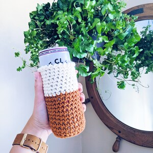 Op de afbeelding: Een gehaakte drankencozy in wit en bruin met een gespikkeld patroon. De cozy zit op een blikje met de zichtbare tekst "CLA".