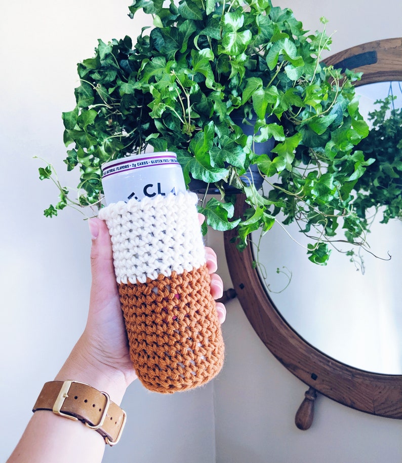 Op de afbeelding: Een gehaakte drankencozy in wit en bruin met een gespikkeld patroon. De cozy zit op een blikje met de zichtbare tekst "CLA".