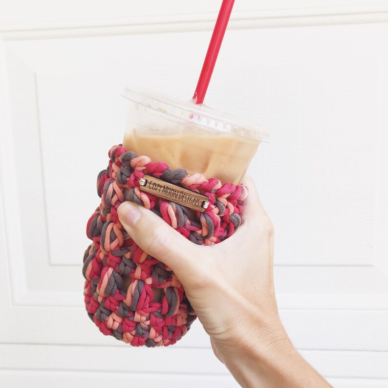 Op de afbeelding: Een gehaakte koffiebeker cozy in roze, rood en bruin met de tekst "Loft Moon Designs" op een houten label. De cozy zit op een doorzichtige plastic beker met ijskoffie.