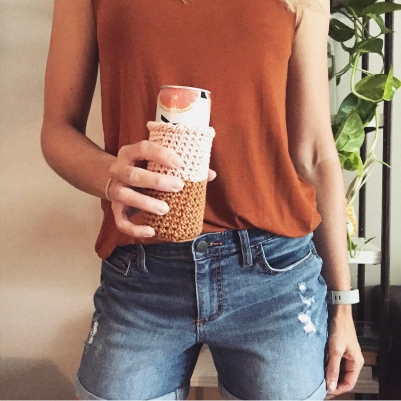 Op de afbeelding: Een persoon houdt een roze en bruine gehaakte blikjeshoes. De hoes zit op een blikje grapefruitsoda.
