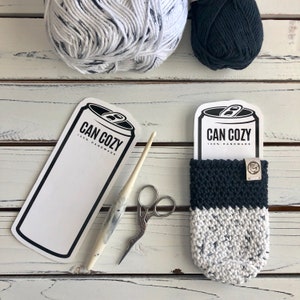 Op de afbeelding: Een zwart-witte gehaakte blikjeswarmer met de tekst "Can Cozy 100% Handmade" op het label. De blikjeswarmer ligt op een wit houten oppervlak met garen en haaknaalden.