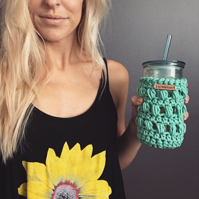Op de afbeelding: Een vrouw houdt een groene gehaakte bekerhoes vast met een bruin label waarop "Cozy Moon Designs" staat, op een heldere glazen beker met een rietje.