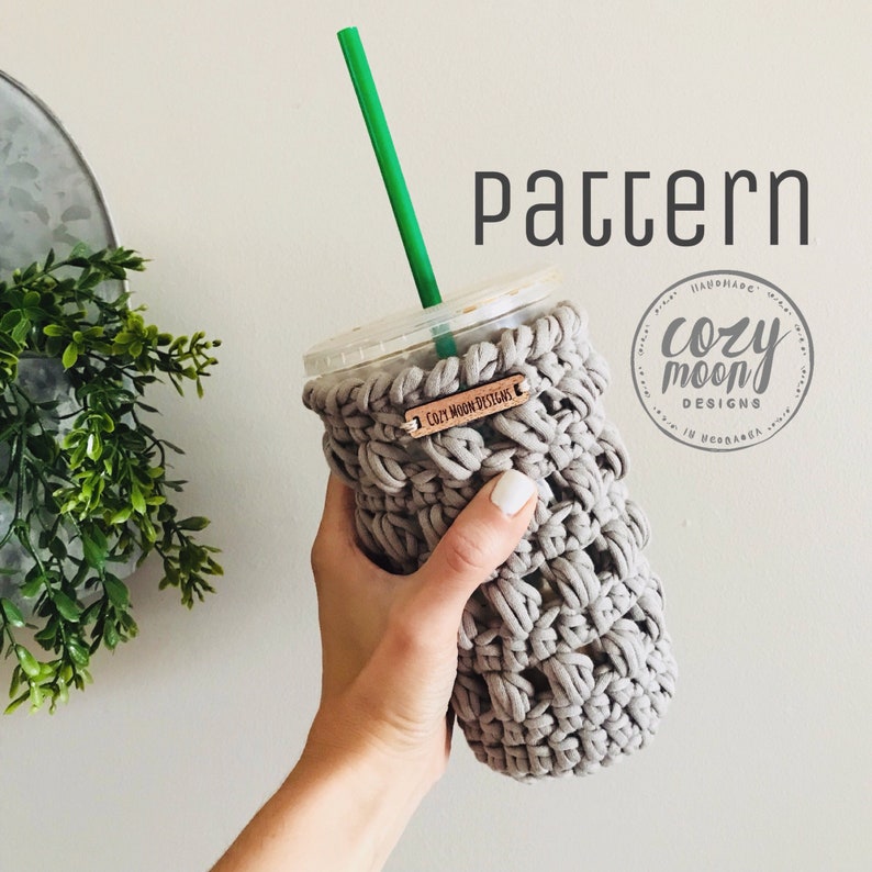 Op de afbeelding: Grijze gehaakte bekerwarmer met een houten label waarop "Cozy Moon Designs" staat.
