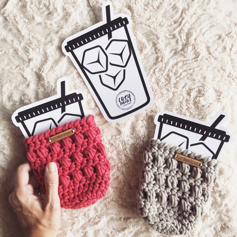 Op de afbeelding: Drie gehaakte bekerwarmers in grijs en roze. Elke bekerwarmer heeft een zwart-witte illustratie van een kopje ijskoffie met de tekst "Cozy Morn Designs".