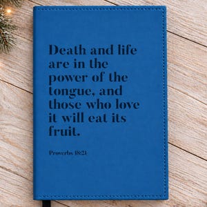 May include: A rectangular blue journal with a black ribbon bookmark. The front cover features the black printed text: Death and life are in the power of the tongue, and those who love it will eat its fruit. Proverbs 18:21. The cover has stitched edges.
