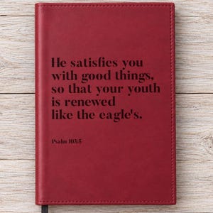 May include: A deep red notebook with a smooth cover featuring the black printed text: He satisfies you with good things, so that your youth is renewed like the eagle's. Psalm 103:5. The rectangular journal includes a small black ribbon bookmark.