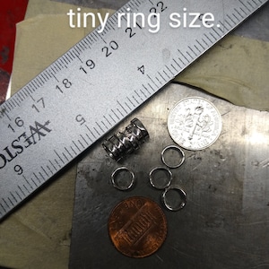 May include: A selection of tiny silver rings, a metal ruler, a stack of metal cylinders, and two coins are shown on a metal surface. The text "Please note the tiny ring size" is visible.