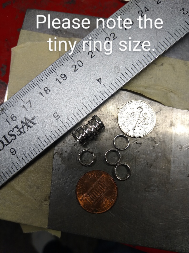 May include: A selection of tiny silver rings, a metal ruler, a stack of metal cylinders, and two coins are shown on a metal surface. The text "Please note the tiny ring size" is visible.
