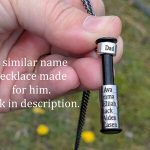 May include: A personalised necklace with a black cylindrical pendant and silver name tags. The top tag reads "Dad", and the lower tags display names. The necklace is held against a blurred green background.