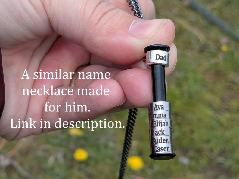 May include: A personalised necklace with a black cylindrical pendant and silver name tags. The top tag reads "Dad", and the lower tags display names. The necklace is held against a blurred green background.