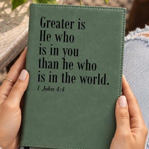 May include: A sage green journal with a smooth, matte finish featuring black text that reads: Greater is He who is in you than he who is in the world. 1 John 4:4. The rectangular notebook has stitched edges and a small black ribbon bookmark.