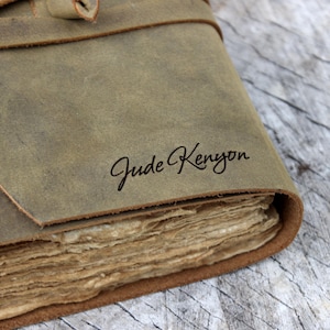 May include: Close-up of a handmade leather journal with a distressed, olive-green cover. The journal features a flap closure and a stack of aged, cream-colored pages. The name "Jude Kenyon" is elegantly scripted on the cover.