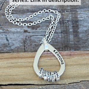 May include: Silver pendant necklace with a teardrop-shaped pendant and chain. The pendant is engraved with the name "KENYONS" and has small rings with names. The image includes the text "Another necklace in this series. Link in description."