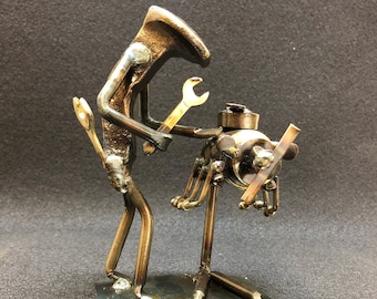 May include: A metal sculpture of a figure holding a wrench and working on a smaller figure. The sculpture is made of recycled metal parts and has a rustic, industrial look.