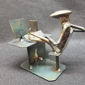 May include: A metal sculpture of a person sitting at a desk with a laptop. The sculpture is made from recycled metal and has a rustic, industrial look.