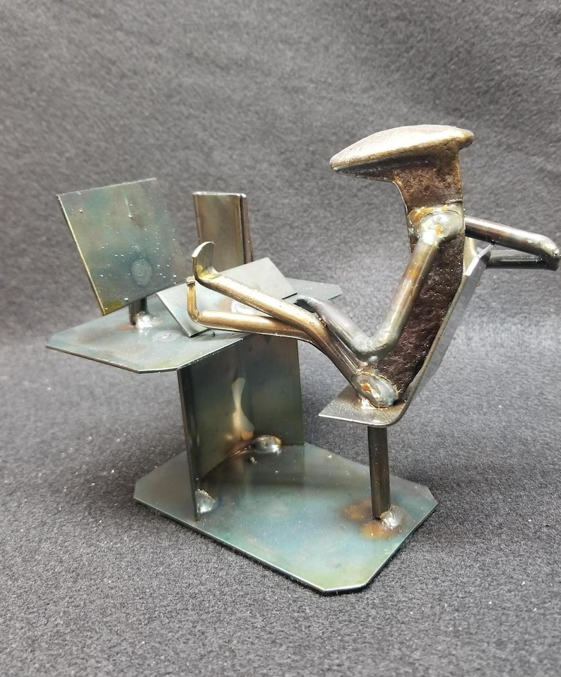 May include: A metal sculpture of a person sitting at a desk with a laptop. The sculpture is made from recycled metal and has a rustic, industrial look.