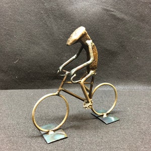 May include: A metal sculpture of a person riding a bicycle. The sculpture is made from recycled metal and is painted gold. The bicycle has two wheels and a seat. The sculpture is standing on two metal bases.