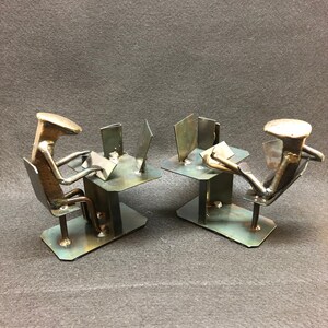 Puede incluir: Dos esculturas metálicas de personas sentadas en escritorios con portátiles. Las esculturas están hechas de clavos y otros restos de metal. Las esculturas están sobre un fondo gris.