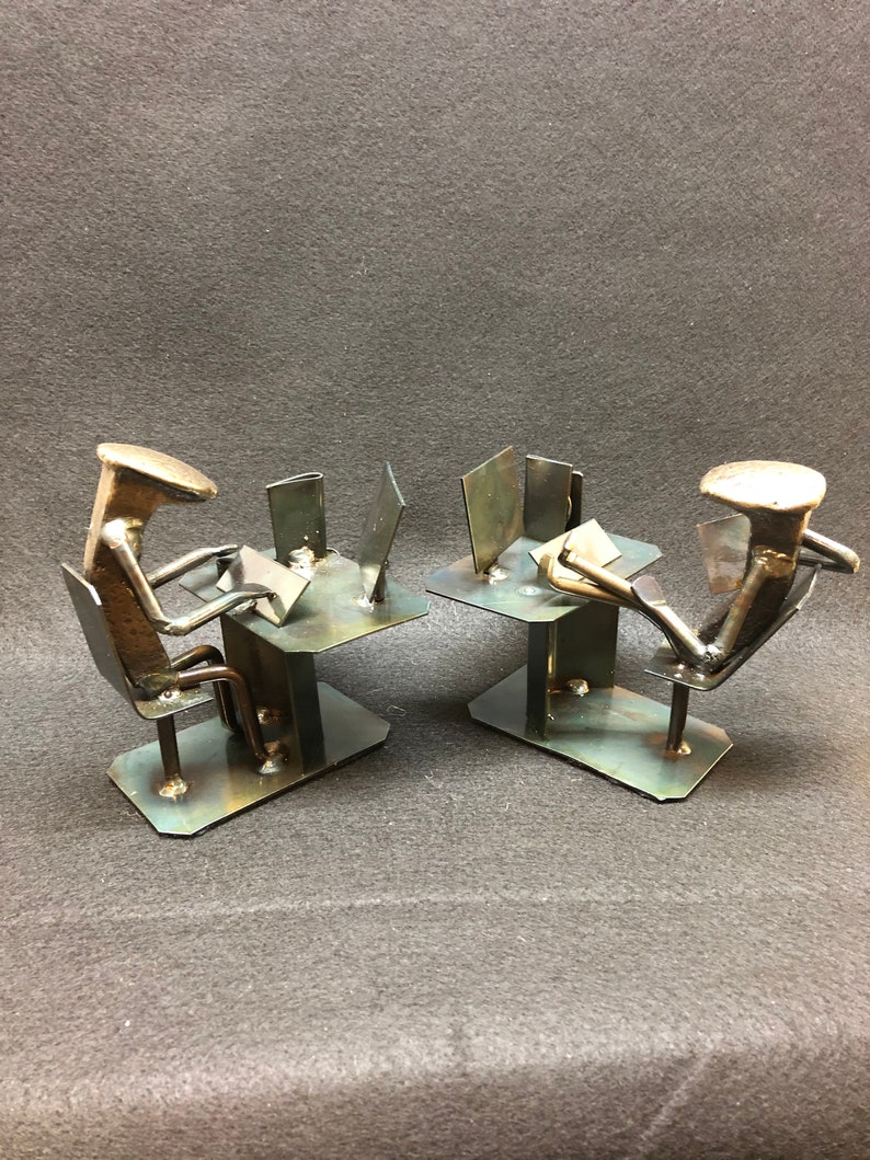 May include: Two metal sculptures of people sitting at desks with laptops. The sculptures are made of nails and other metal scraps. The sculptures are on a grey background.