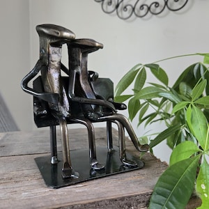 May include: A metal sculpture of two figures sitting on a bench. The figures have a dark, aged metal finish. The bench is also metal, and the sculpture is mounted on a rectangular base. The sculpture is placed on a wooden surface. The sculpture is approximately 15 cm in height.