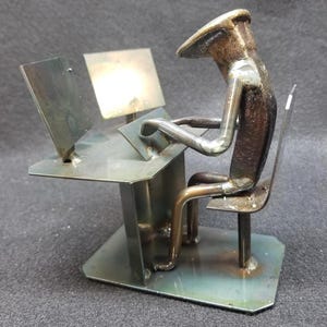 May include: A metal sculpture of a person sitting at a desk with a computer monitor and keyboard. The sculpture is made from recycled metal and is painted in a metallic silver finish.