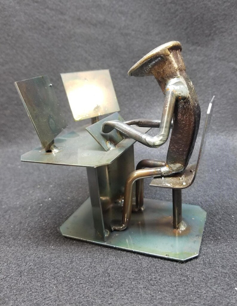 May include: A metal sculpture of a person sitting at a desk with a computer monitor and keyboard. The sculpture is made from recycled metal and is painted in a metallic silver finish.