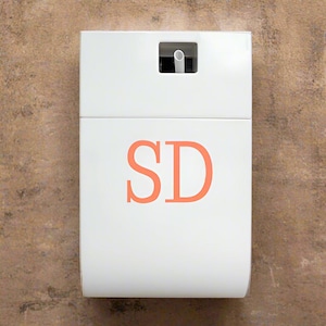 Puede incluir: Un portarrollos de papel higiénico blanco, rectangular, montado en la pared, con una base curva. El soporte presenta las letras color coral "SD" en la parte delantera. Una pequeña abertura en la parte superior revela una parte del rollo de papel higiénico. El fondo es una superficie marrón texturizada.