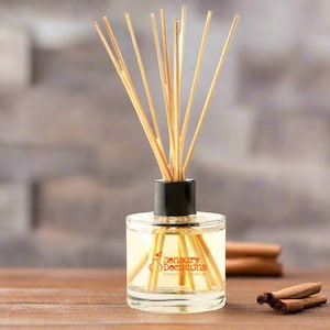 Puede incluir: Una botella de difusor de vidrio transparente con una tapa negra y palitos de madera. La botella está llena de un líquido amarillo claro y tiene una etiqueta que dice "Sensory Decisions Unique Fragrances". Hay palitos de canela en la mesa junto al difusor.