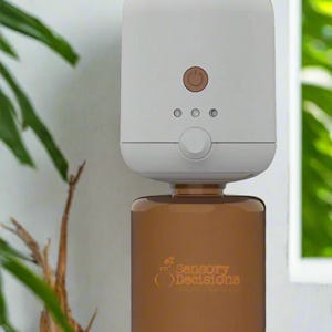 Op de afbeelding: Witte, wandgemonteerde diffuser met een bruine glazen fles gevuld met etherische olie. De diffuser heeft een aan/uit-knop en een knop om de instellingen aan te passen. De fles heeft de tekst "Sensory Decisions" erop gedrukt.