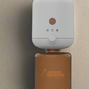 Può includere: Diffusore di oli essenziali elettrico bianco con una bottiglia di vetro marrone piena di olio essenziale. La bottiglia ha il logo "Sensory Decisions" stampato su di essa.