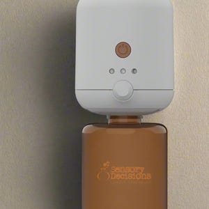 Puede incluir: Ambientador automático blanco y marrón con el texto "Sensory Decisions" en naranja. El dispositivo tiene una parte superior blanca con un botón de encendido y tres luces indicadoras. La parte inferior marrón contiene la fragancia.
