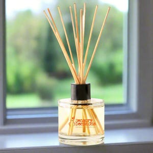 Puede incluir: Un difusor de aromaterapia de vidrio transparente con una tapa negra y varillas de madera. La botella contiene un líquido amarillo claro y presenta el texto "Sensory Decisions Unique Fragrances" en naranja. El difusor está colocado frente a una ventana con un fondo verde borroso.
