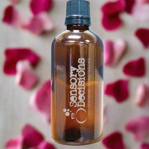 Puede incluir: Una botella de vidrio marrón con tapa negra, con el texto "Sensory Decisions Unique Fragrances". La botella está rodeada de pétalos de rosa borrosos rosas y rojos, creando un fondo suave y romántico.