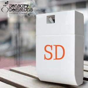 May include: A white rectangular fragrance diffuser with the orange letters "SD" on the front. The top has a small opening with a white tube. The logo "Sensory Decisions, Unique. Fragrances." is in the upper left corner. Smoke is visible above the diffuser.