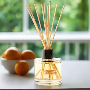 May include: A glass diffuser bottle with a black cap and wooden reeds. The bottle contains a yellow liquid and the words "Sensory Decisions" are printed on the front. Oranges in a white bowl are in the background.