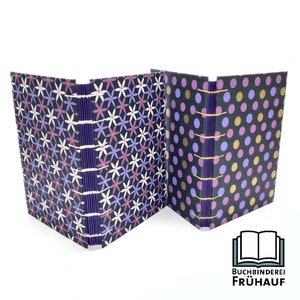 May include: Two handmade journals with purple covers. One journal has a floral pattern and the other has a polka dot pattern. Both journals have a purple spine with gold stitching.