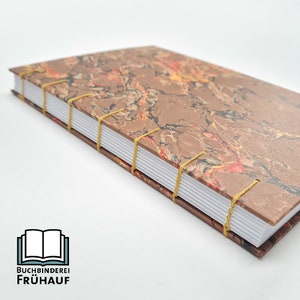 May include: A brown marbled notebook with gold stitching and a white spine. The notebook is closed and the spine is facing the camera. The notebook is on a white surface.