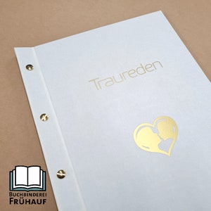 Könnte beinhalten: Ein weißes Buch mit goldenem Text "Traureden" und einem goldenen herzförmigen Design mit zwei Silhouetten. Das Buch ist mit goldenen Nieten gebunden. Das Logo von "Buchbinderei Frühauf" befindet sich in der unteren linken Ecke.