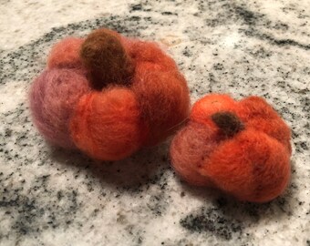 May include: Two handmade felt pumpkins in shades of orange, red, and purple. The pumpkins have brown stems and a textured, fuzzy appearance. One pumpkin is larger than the other, suggesting a fall or harvest theme.