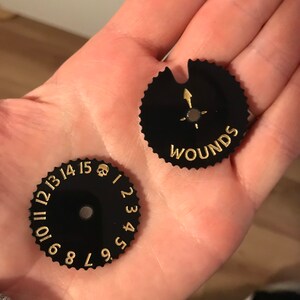 May include: Two black circular counters with gold numbers and a skull symbol. One counter has the word "WOUNDS" printed on it.