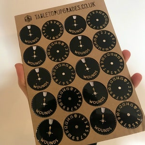 May include: A sheet of brown cardstock with 12 black and gold circular wound trackers. Each tracker has a gold skull and the numbers 1 through 15 around the edge.