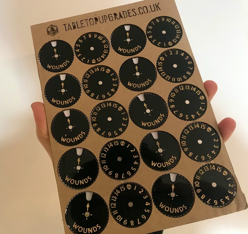 May include: A sheet of brown cardstock with 12 black and gold circular wound trackers. Each tracker has a gold skull and the numbers 1 through 15 around the edge.