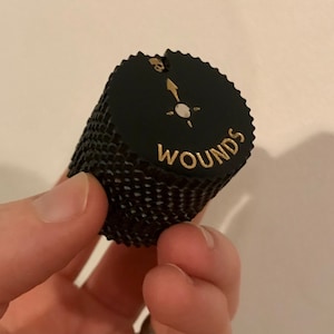 May include: Black and gold dice with the word "Wounds" printed on the side. The dice has a skull and crossbones symbol on the top.