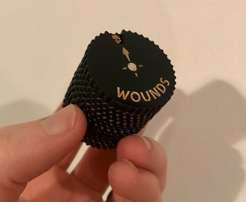 May include: Black and gold dice with the word "Wounds" printed on the side. The dice has a skull and crossbones symbol on the top.