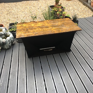 May include: A black wooden beehive with a golden top and a small metal handle. The beehive is sitting on a grey wooden deck.