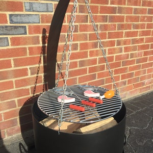 May include: A black metal fire pit with a hanging grill grate. The grill grate is suspended by chains and has hot dogs and pink meat on it. The fire pit is sitting on a brick patio.