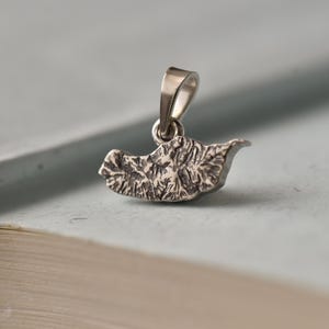 Puede incluir: Un pequeño colgante de plata con un diseño de hoja texturizada. El colgante tiene un lazo en la parte superior para una cadena y se muestra sobre una superficie azul claro. El diseño de la hoja tiene detalles intrincados.